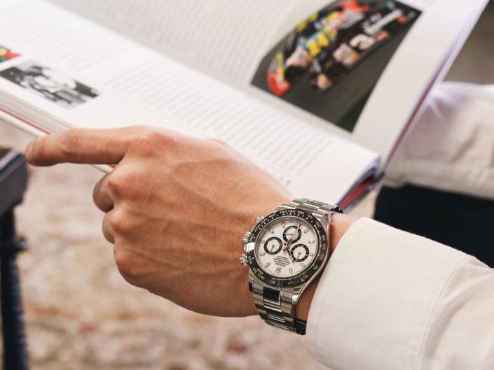 A man wearing a Rolex Daytona watch reading a book about race cars